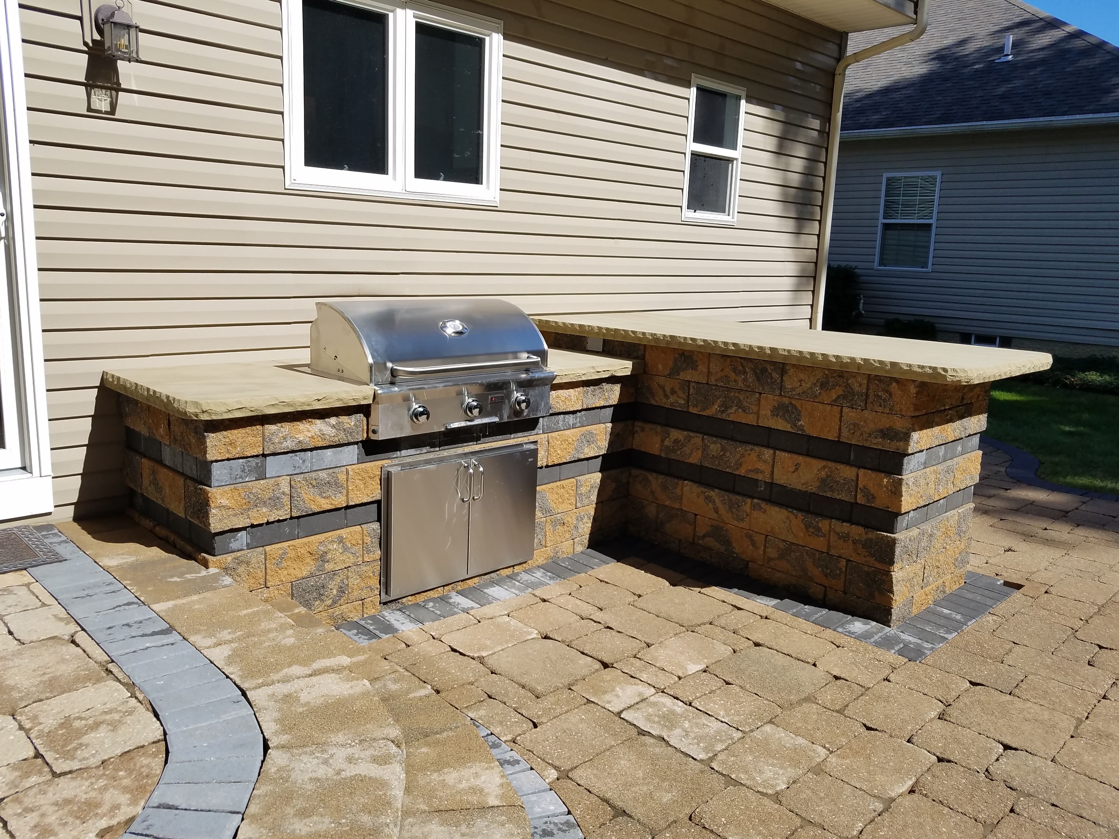 Stainless steel grill built into a tan stone block outdoor kitchen on a paved patio.