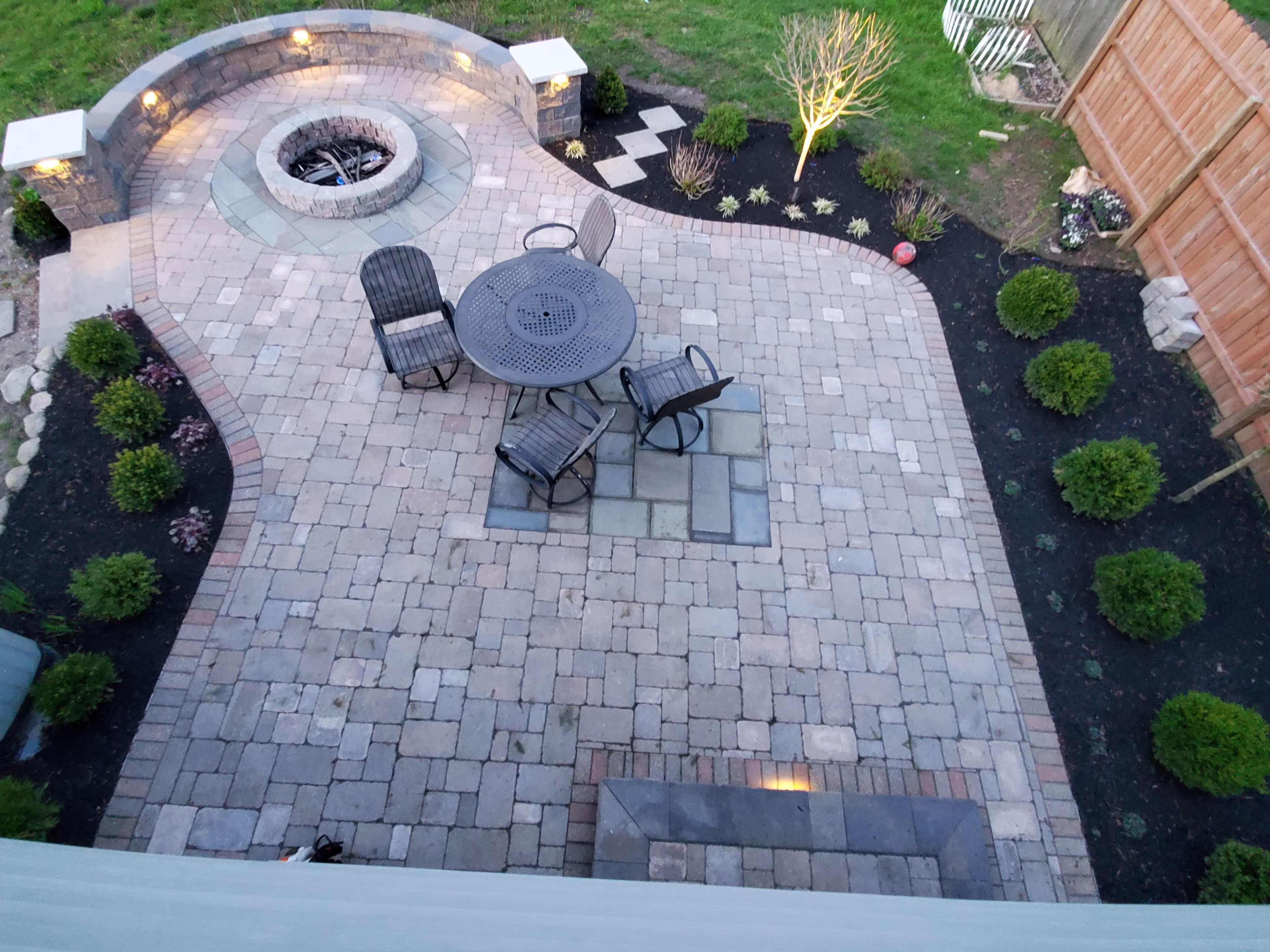 Stone paver patio featuring a circular fire pit, dining set, and illuminated garden beds.