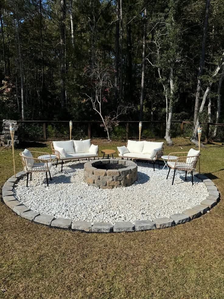 Circular stone fire pit on white gravel with white outdoor seating in a wooded backyard.