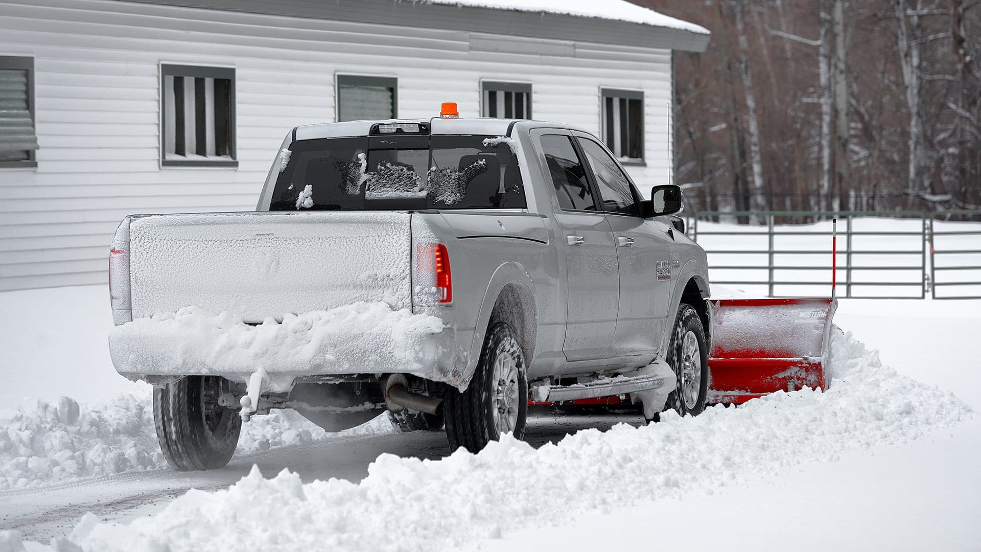 Silver pickup truck using a red plow to clear heavy snow from a residential driveway.