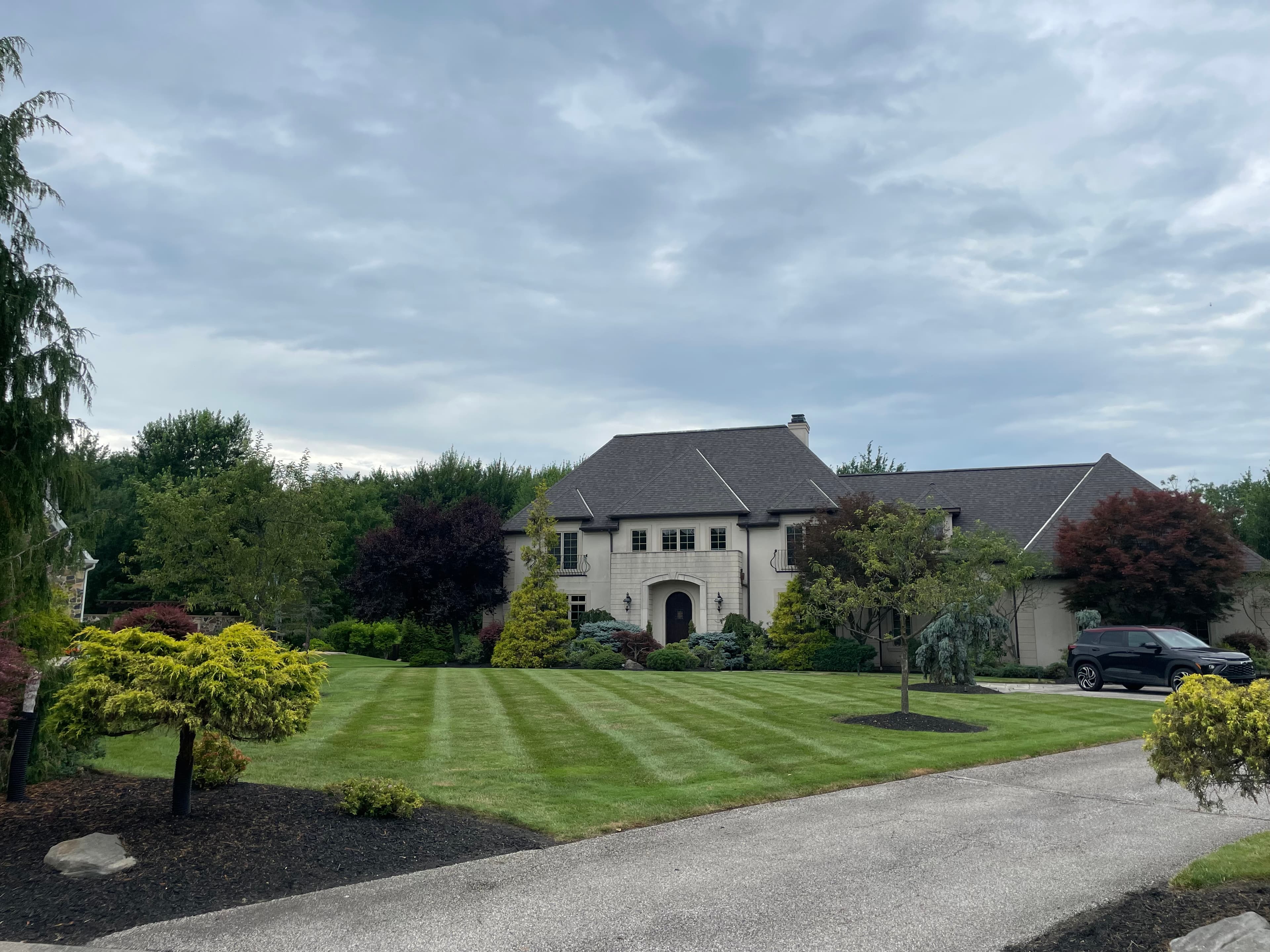 Large stone house with a perfectly striped lawn and lush landscaping under cloudy skies.